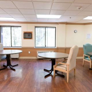 An empty room with wooden floors, tables, and chairs near large windows.