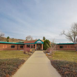 A brick building with a green roof and a pathway leading to the entrance, surrounded by landscaping and trees under a blue sky.