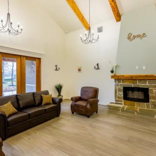 A spacious living room featuring a cozy sofa and armchair, a stone fireplace, and elegant chandeliers under wooden ceiling beams.