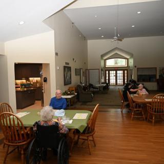 A community dining area where elderly individuals are seated at tables, some in wheelchairs, enjoying a meal.