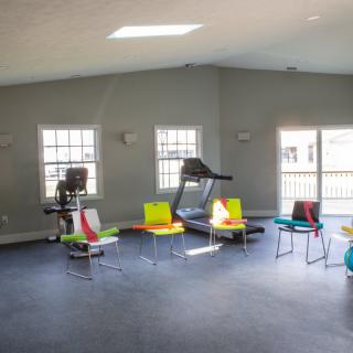 A small gym with exercise equipment and colorful chairs by the windows.