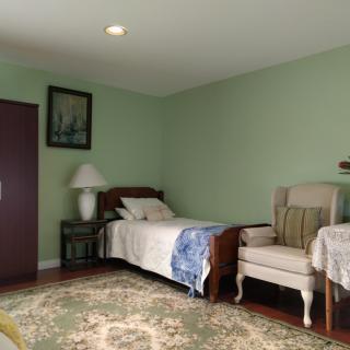 A cozy bedroom with green walls, featuring a single bed, an upholstered chair, and a side table with a lamp. A piece of artwork hangs on the wall, and an ornate rug covers the floor.