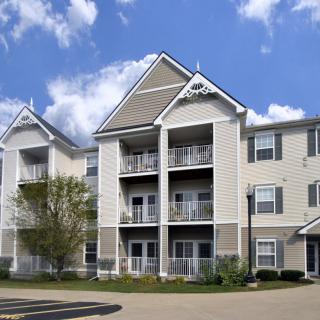 A modern three-story apartment building with multiple balconies and a well-maintained exterior, set against a blue sky with clouds.