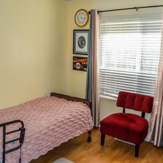 A cozy bedroom with a single bed, red chair, and light streaming in through a window.