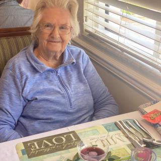 An elderly woman sitting at a table in a restaurant, smiling with daylight shining through the window.