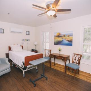 A cozy and well-lit hospital room featuring a bed, a small table with chairs, and a ceiling fan.