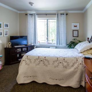 A cozy bedroom with a neatly made bed, television, and window with sheer curtains.