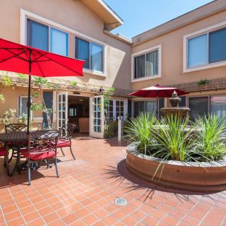 A sunny courtyard with patio furniture and red umbrellas, surrounded by windows.