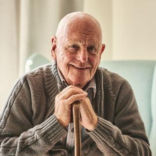 An elderly man smiling warmly indoors, resting his hands on a cane.