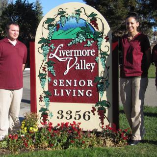 Two people standing next to a Livermore Valley Senior Living sign outdoors.