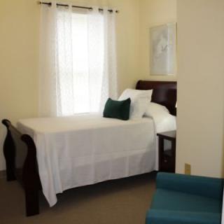 A cozy bedroom featuring a bed with white linens and decorative pillows, next to a window with sheer curtains.