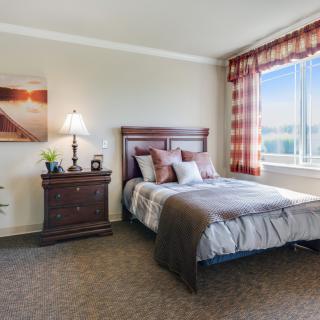 A cozy bedroom with a neatly made bed, nightstand, lamp, and a window providing natural light.