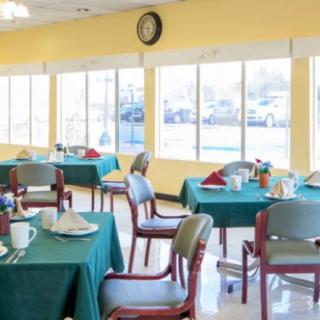 A bright dining area with tables set for a meal, featuring large windows and a clock on the wall.