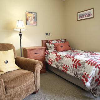 A cozy bedroom with a single bed and an armchair, complemented by a lamp and decorative pillows.