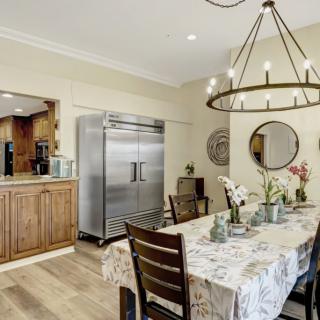 A cozy dining room with a large table, wooden chairs, a chandelier, and a modern refrigerator in the background.