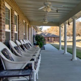 A row of white wicker chairs lined up on a covered porch with ceiling fans overhead.