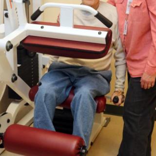 An elderly person receiving assistance with gym equipment.
