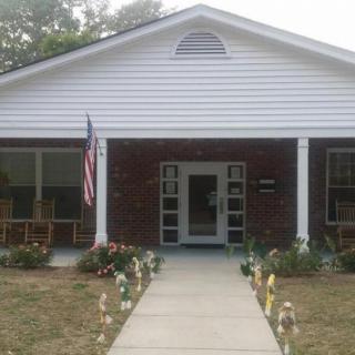 A brick building with a porch and an American flag on display, surrounded by a garden.