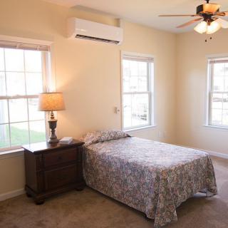 A cozy bedroom with a single bed, nightstand, and lamp, featuring an air conditioner and ceiling fan.