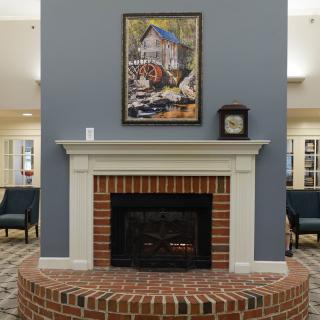 A cozy room featuring a brick fireplace with a painting and clock on the mantel, surrounded by comfortable chairs on a patterned carpet.