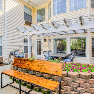 A cozy outdoor patio with a wooden bench, pergola, and seating area surrounded by plants.