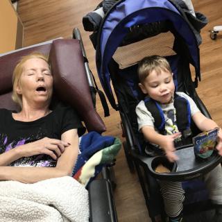An elderly person resting in a chair beside a child sitting in a stroller indoors.