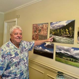 A man in a floral shirt is smiling and pointing at paintings on a wall indoors.
