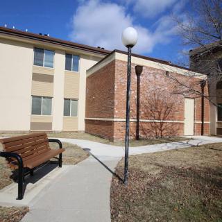 A courtyard with a bench and lamppost alongside a brick and stucco building under a partly cloudy sky.