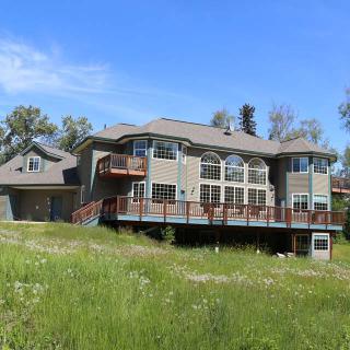 A large modern house with a spacious deck, surrounded by greenery and under a clear blue sky.