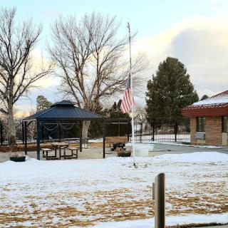 A winter scene featuring a gazebo in a park, surrounded by snow and trees, with an American flag standing tall.
