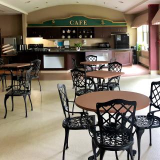 An empty cafe interior featuring several round tables and ornate chairs, with a kitchen area in the background.