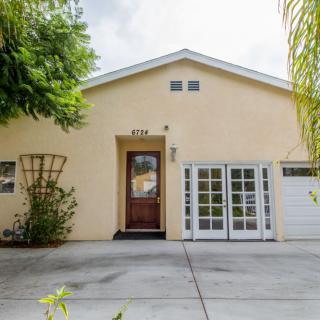 The image shows the exterior of a beige-colored house with a driveway, wooden front door, and white garage doors.