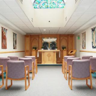 A small chapel interior with seating facing an altar and stained glass window above.