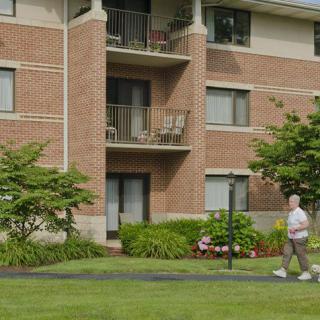 A person walking past a brick apartment building with balconies, surrounded by a well-maintained lawn and trees.