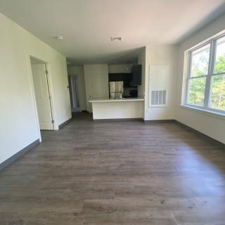 Spacious empty living room with wooden floors and a view into the kitchen, illuminated by natural light from a large window.