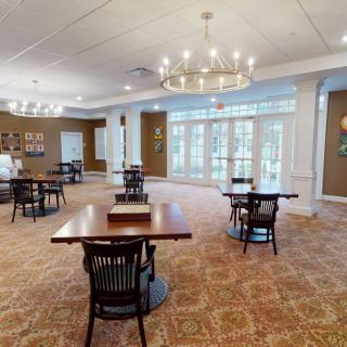 A spacious dining area with elegant chandeliers, wooden tables, and chairs arranged on a patterned carpet, complemented by large windows for natural light.