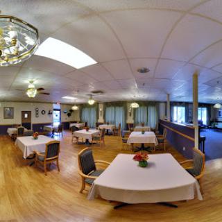 A spacious and elegantly arranged dining room featuring tables set for guests, adorned with chandeliers and a warm wooden floor.