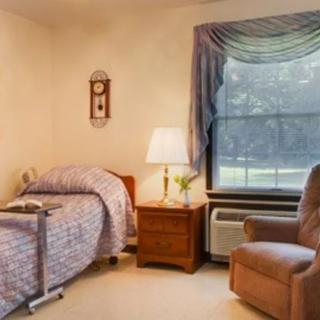 A cozy bedroom with a single bed, bedside table, lamp, and an armchair near a large window.
