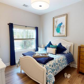 A cozy bedroom featuring a neatly made bed with blue-themed cushions, a framed artwork on the wall, and natural light streaming through a window with dark curtains.