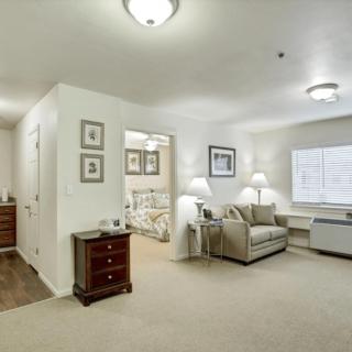 A cozy apartment interior featuring a living area with a sofa, armchair, and a well-lit bedroom.