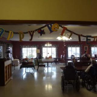 A community room decorated with a festive banner and filled with chairs and tables, welcoming natural light through large windows.