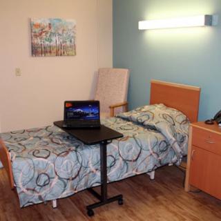 A small hospital room with a bed featuring a patterned bedspread, a laptop on an overbed table, and simple wooden furniture.