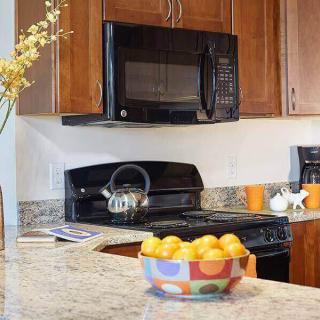 A modern kitchen with wooden cabinets, featuring a microwave, stovetop with a teapot, and a colorful fruit bowl filled with lemons on the countertop.