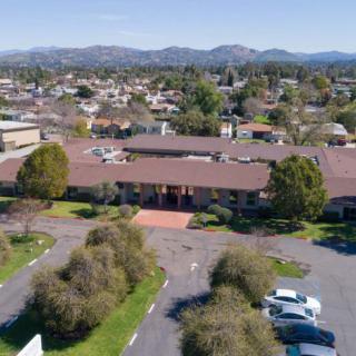 Aerial view of a suburban area with a central building, surrounded by trees and parked cars, with mountains in the background.