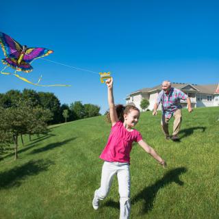 A child and an adult flying a kite on a grassy lawn under a clear blue sky.