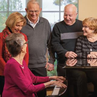 A group of seniors gathered around a piano, enjoying music and each other's company indoors.