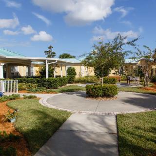 A beautifully landscaped garden with a circular pathway and surrounding buildings under a blue sky with clouds.