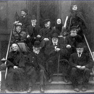 A historical black and white photo of a group of people in formal attire sitting on a staircase.