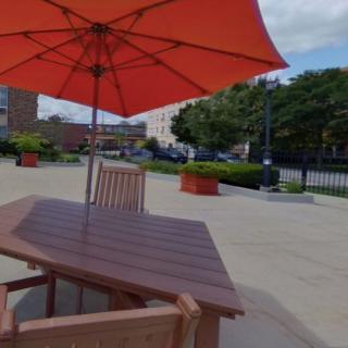 An outdoor patio area with a wooden table and chairs under an orange umbrella, set in an urban environment.