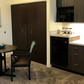 A modern kitchenette setup with a small table and chair, featuring cabinets, a microwave, and a mini fridge.